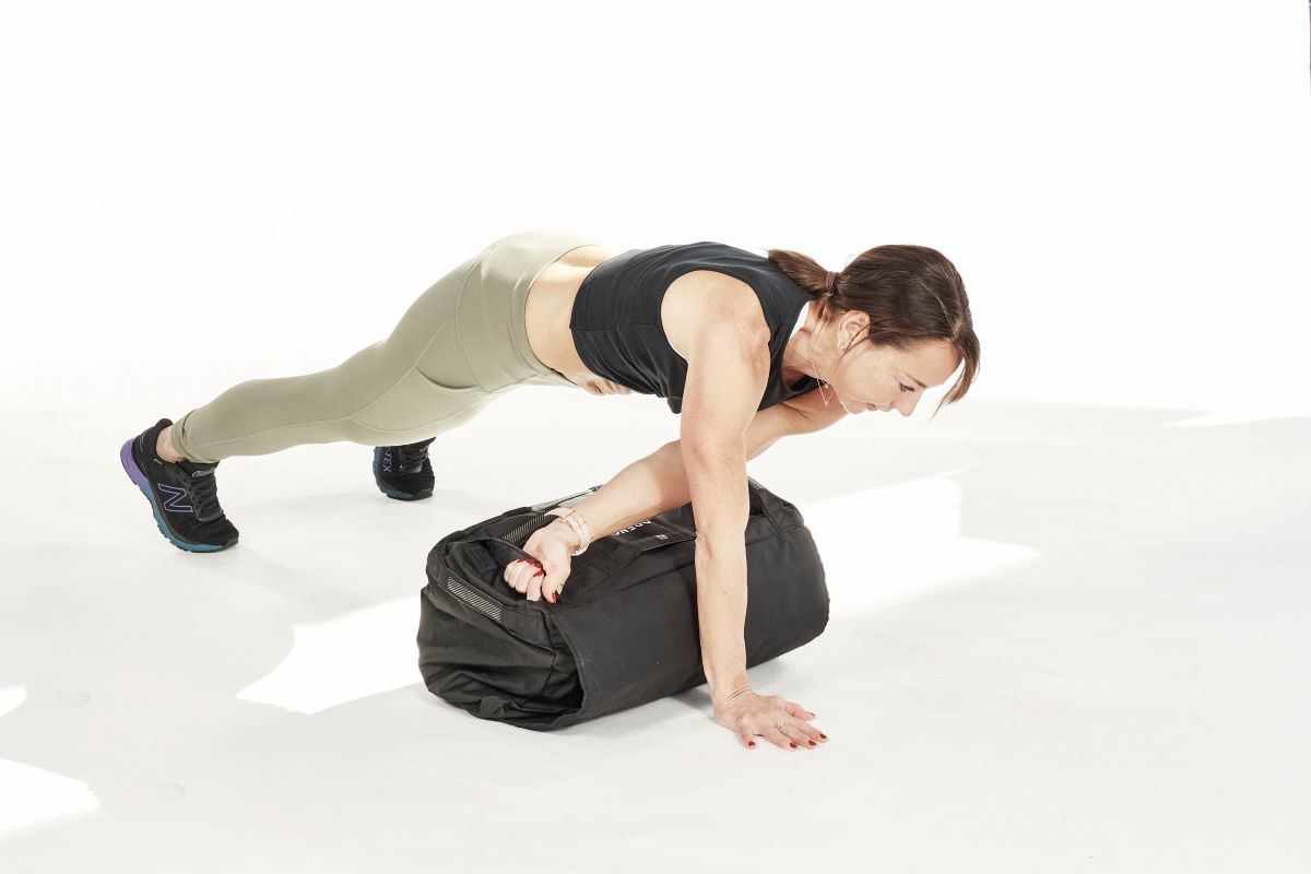 woman demonstrates plank lateral drag woman demonstrates thrusters, one of the best sandbag exercises