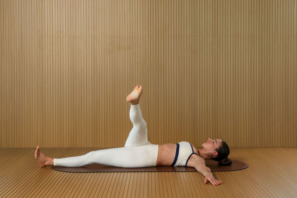 woman performs Pilates lymphatic drainage workout, she is lying on her back with arms outstretched circling her right leg