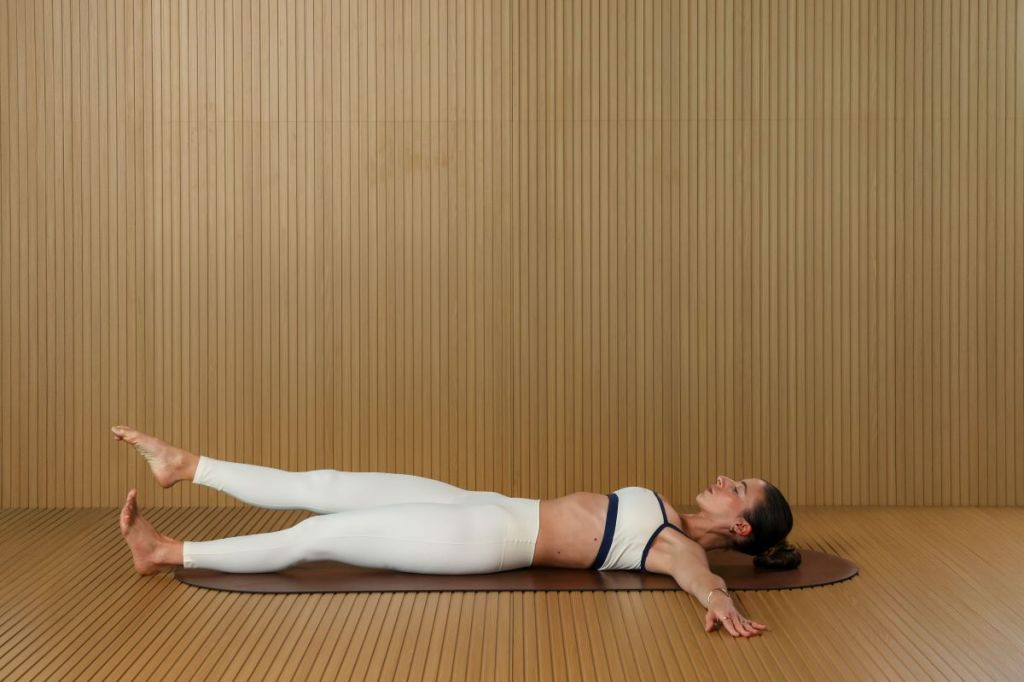 woman performs Pilates leg circles for lymphatic drainage, she is lying on her back with arms outstretched circling her right leg