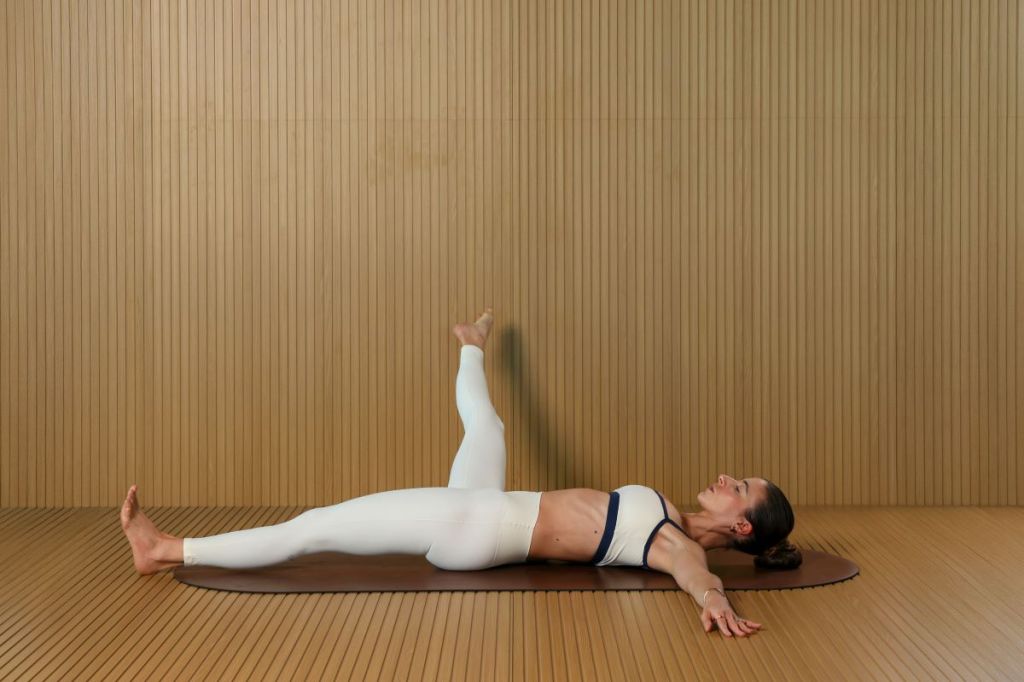 woman performs Pilates leg circles for lymphatic drainage, she is lying on her back with arms outstretched circling her right leg out to the side
