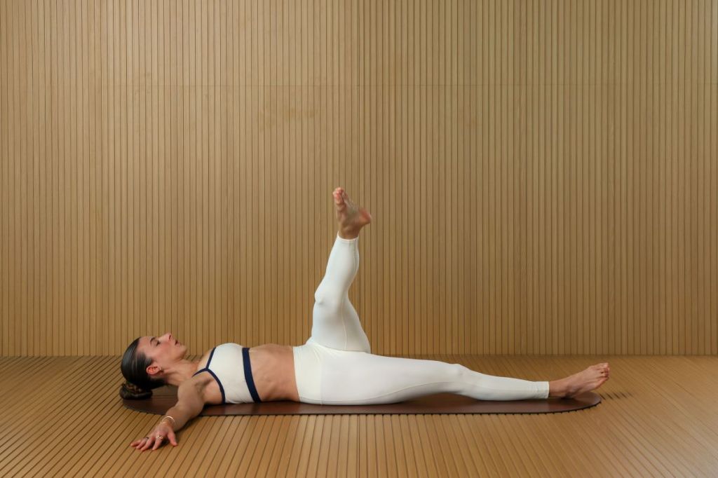 woman performs Pilates lymphatic drainage workout, she is lying on her back with arms outstretched circling her left leg