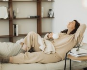 Young woman wearing cozy costume sitting on grey sofa couch and drinking tea. Woman spending free time in living room, enjoying morning, being in dreamy and relaxed mood. People lounge concept.
