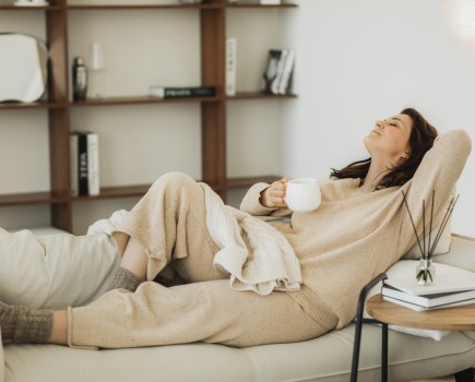 Young woman wearing cozy costume sitting on grey sofa couch and drinking tea. Woman spending free time in living room, enjoying morning, being in dreamy and relaxed mood. People lounge concept.