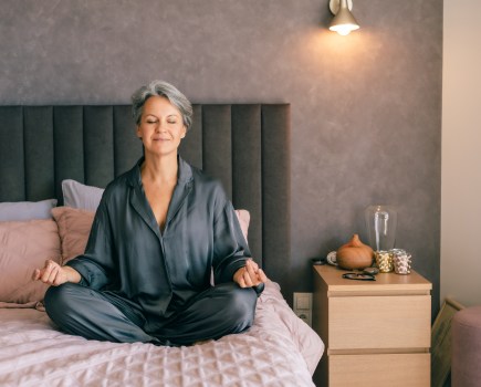 A mature woman with gray hair sits in a meditative pose on her bed, eyes closed. The peaceful moment reflects midlife balance, menopause self-care, and emotional grounding.